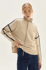 Sweater de tejido de punto color beige, con cuello redondo y detalles de bordado en zigzag en color azul oscuro a lo largo de las mangas y hombros. Presenta un pequeño bordado con texto en el pecho.