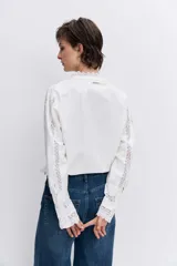 Camisa de algodón color blanco con detalles de encaje broderie y alforzas verticales. Presenta cuello alto con volados, cierre frontal de botones y mangas largas con puños acampanados.