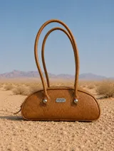 Cartera estilo baguette de cuero color tostado con textura grabada. Presenta una silueta alargada y estructurada, con manijas largas y detalles de nudos esféricos metálicos en la unión de las mismas. Incluye placa metálica con el logo de la marca en el frente.