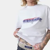 Camiseta blanca de corte "boxy fit" con estampado frontal que dice "emestudios" en azul con detalles rojos, y debajo "EXCLUSIVE PIECES SELECTED WITH CARE / MADRID".