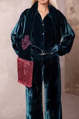 Camisa de seda en color verde petróleo, con corte recto y estructura clásica. Presenta un diseño con apliques bordados en tono borravino y detalles de flecos largos en la parte frontal inferior.