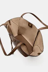 Bolso de hombro tipo maxi shopper color marrón, con doble asa y estructura flexible que se pliega hacia afuera. Presenta un detalle de tira ajustable con cordón en el centro frontal. Incluye un compartimento interior extraíble con cierre de cremallera y cierre principal mediante botón imantado.