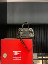 Cartera de mano y hombro color beige, de material símil cuero, con forma estructurada y redondeada. Posee dos asas cortas superiores y una correa larga ajustable y desmontable. Cuenta con un bolsillo exterior con cierre en la parte frontal.