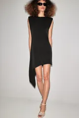 Vestido corto negro sin mangas con hombreras, cuello redondo y bajo asimétrico con una caída lateral más larga. Cuenta con cierre en la espalda con abertura y botón.