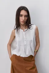 Blusa sin mangas de color blanco, confeccionada en tejido fluido con cuello redondo y detalle de alforzas verticales en el frente. Cuenta con una abotonadura parcial en el centro.