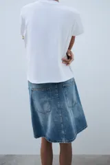 Falda de tiro medio confeccionada en denim, con trabillas, cinco bolsillos, cierre cruzado con botones y bajo acabado asimétrico.