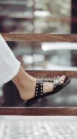 Sandalias planas de cuero negro, con diseño de tiras anchas interconectadas y punta cuadrada, decoradas con tachas metálicas doradas.