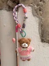 Llavero con dije de oso de peluche marrón, con gafas redondas, suéter de punto rosa y blanco con perlas y falda plisada a rayas. El oso sostiene un globo rosa. El colgante es una correa de cuerda trenzada multicolor con detalles en rosa neón.
