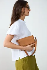 Cartera de hombro pequeña de cuero tostado, con solapa frontal decorada con bordados artesanales en los bordes y cierre de broche magnético. Incluye asa corta de mano y forro interior.