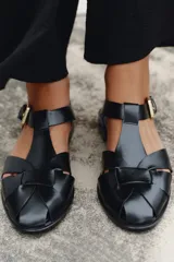 Sandalia plana tipo cangrejera de color negro, con diseño de tiras cruzadas en el empeine y cierre mediante hebilla metálica dorada en el lateral.