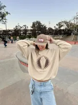 Buzo de cuello redondo color tostado, con estampa frontal de un esqueleto dentro de un ataúd rodeado por un diseño solar y la inscripción 'Wake Up' junto a 'Kaboa Industries'.