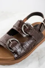 Sandalias bajas color marrón con textura croco, dos tiras anchas sobre el empeine con hebillas metálicas plateadas y tira trasera ajustable con hebilla.