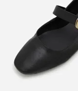 Zapatilla tipo bailarina de cuero negro, con punta redondeada, tacón bajo y una tira decorativa en el empeine con hebilla metálica dorada.