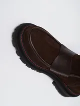 Botineta de gamuza marrón con detalles en cuero liso al tono. Presenta una suela track negra con costuras en contraste color rojo y una tira vertical roja en el talón.
