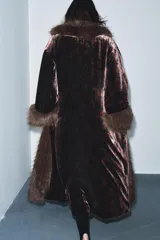 Abrigo largo de terciopelo en tono marrón oscuro, con cuello solapa y puños adornados con aplicación de pelo sintético. Presenta un diseño abierto con forro interior en color rojo con estampado floral.