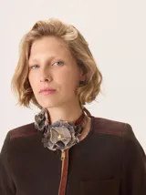 Chaqueta de paño color marrón oscuro, de corte clásico y cuello redondo, con cierre frontal dorado. Presenta bolsillos rectangulares con ribetes en color rojizo y parches de pana a rayas en los hombros.