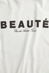 Remera de algodón color blanco crudo, con cuello redondo y manga corta. Presenta un estampado frontal con la palabra "BEAUTÉ" en mayúsculas y debajo en cursiva "Rue des Artistes France".