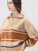 Pullover de textura tipo terciopelo con cuello alto y cierre frontal parcial. Presenta un diseño a rayas horizontales en tonos grises y negros, con detalles en rojo y blanco en el cuello, puños y ruedo. Incluye un aplique bordado con la letra M en el pecho y puños acanalados.