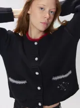 Cardigan negro de algodón grueso con cuello redondo y cierre frontal de botones joya. Presenta bolsillos frontales con detalles de pelo sintético gris y logo bordado en uno de ellos. Posee puños y ruedo elastizados.