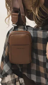 Bandolera pequeña de diseño compacto en color mostaza, confeccionada en material sintético con textura granulada. Presenta un bolsillo frontal con el logo de la marca impreso, cierre superior y correa ancha ajustable y desmontable.