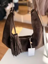 Cartera tipo hobo de textura gamuzada en color marrón oscuro, con diseño flexible y asa larga regulable. Presenta un detalle de flecos largos desmontables en uno de los laterales y cierre superior.