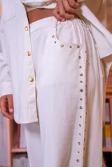 Pantalón de denim blanco de corte amplio, con cierre de botón y cremallera, y detalles de tachas en los laterales y bolsillos.