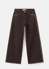 Pantalón de jean color marrón oscuro, de corte recto y tiro medio. Presenta una hilera de tachas metálicas plateadas a lo largo de la costura lateral externa.