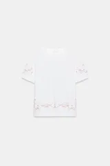 Camiseta de algodón de corte regular, color blanco, con cuello redondo y manga corta. Presenta detalles de bordados florales y ornamentales en color rosado en el ruedo y en las mangas.