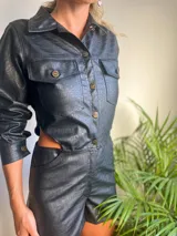 Mono corto negro de cuero sintético con textura croco, cuello camisero, cierre frontal con botones dorados, mangas largas con puños abotonados, bolsillos con solapa en el pecho y aberturas laterales en la cintura.
