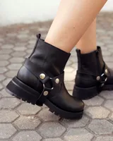 Bota de caña alta confeccionada en cuero negro, con suela track de goma. Presenta un diseño versátil con dos hebillas ajustables en la parte superior de la caña y una correa decorativa con tachas metálicas y argollas en la zona del tobillo.