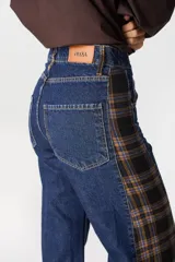 Jean de pierna ancha con diseño bicolor, combinando denim azul clásico con un panel lateral de tela a cuadros en tonos marrón y negro. Presenta cuatro bolsillos funcionales y cierre frontal.