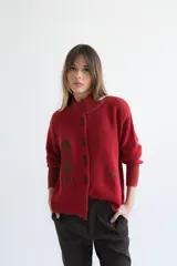 Cardigan de punto color rojo con cuello alto asimétrico, cierre frontal de botones oscuros y detalles de estampado geométrico en contraste. Posee mangas largas con puños acanalados anchos.