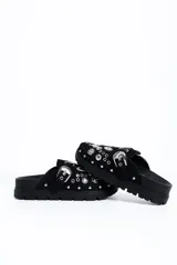 Zueco negro de estilo slip-on, confeccionado con acabado similar a gamuza, suela gruesa dentada de goma, hebilla lateral plateada de diseño occidental y apliques metálicos decorativos con tachas y ojetes en la capellada.