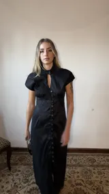 Vestido largo negro de tafeta, con silueta entallada, cuello alto con abertura en forma de gota en el escote, mangas cortas y hilera de botones decorativos en el frente.