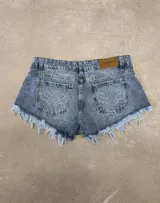 Short de denim azul con efecto deslavado, tiro medio y dobladillo desmechado con flecos. Presenta bolsillos traseros con bordado en forma de alas y etiqueta de cuerina de la marca Ask Denim en la pretina.