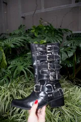 Bota estilo biker de caña alta, confeccionada en cuero vacuno color rojo cherry. Presenta un diseño con múltiples tiras horizontales con hebillas metálicas, punta redondeada y taco cuadrado bajo de madera.