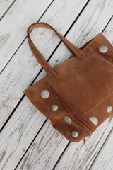 Bolso estilo shopper de gamuza color marrón, con diseño artesanal que incluye flecos laterales y apliques de tachas metálicas circulares. Cuenta con doble asa de hombro y costuras a la vista.