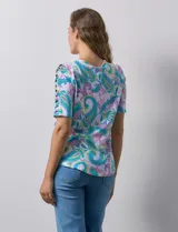 Remera de algodón estampado con diseño paisley en tonos celeste, rosa y amarillo. Tiene cuello redondo y mangas cortas con detalle de entrelazado y aberturas en los hombros.
