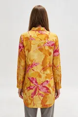 Camisa de lino larga y holgada, de color amarillo con estampado abstracto en tonos fucsia y naranja, con cuello camisero, cierre frontal parcial con botones y aberturas laterales en el dobladillo.