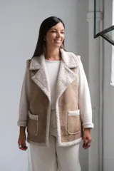 Chaleco de gamuza color beige con interior y detalles de corderito color crudo. Presenta solapas amplias, bolsillos frontales aplicados y diseño sin mangas.