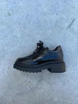 Zapato mocasín negro de cuero charol con diseño tipo reptil, cordones y suela track.
