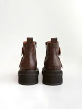 Botas cortas de cuero color marrón con suela track negra. Presentan dos correas ajustables con hebillas metálicas plateadas en el empeine y cierre lateral interno.