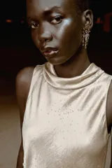 Top satinado color beige con cuello fluido, manga sisa y aplicaciones de brillos. Presenta falsos botones forrados en el lateral, bajo asimétrico con abertura y cierre de cremallera oculta.