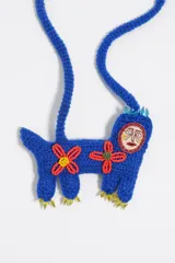 Collar artesanal de crochet en color azul con un colgante en forma de criatura fantástica. El diseño presenta una máscara bordada con rostro, flores decorativas en rojo y amarillo, y detalles de cuentas de vidrio en las extremidades. El cordón es de tejido de punto azul con cierre de nudo ajustable.