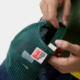 Gorra verde oscura tipo trucker con frente de cuerina, visera plana y paneles traseros de malla. Cuenta con escudo bordado en la parte frontal y cierre snapback ajustable.