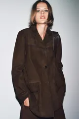 Blazer de mujer color marrón oscuro, confeccionado en piel de ante 100%. Presenta cuello solapa de muescas, manga larga, bolsillos de plastrón frontales y cierre frontal de botones.