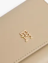 Billetera plegable en tres partes (trifold) de color beige claro, con acabado liso y cierre de solapa. Presenta una insignia metálica dorada con el monograma TH en el frente y un bolsillo exterior con cierre de cremallera.