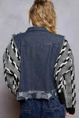 Campera de jean color negro con cuerpo de denim desgastado y mangas tejidas en contraste con diseño geométrico en blanco y negro. Presenta cuello camisero, cierre frontal con botones metálicos y detalles de desflecado en las costuras.