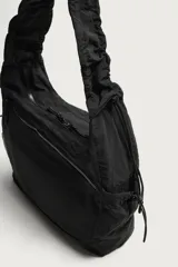Bolso de hombro negro de nylon, con compartimento principal y bolsillos con cierre de cremallera, bolsillos laterales con cordón ajustable y asa fruncida.