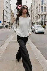 Blusa de crepe con cuello barco y manga larga. Presenta un diseño minimalista en color blanco con detalles de vivos en color negro que recorren el cuello, las mangas y los puños. Los puños incluyen botones decorativos.